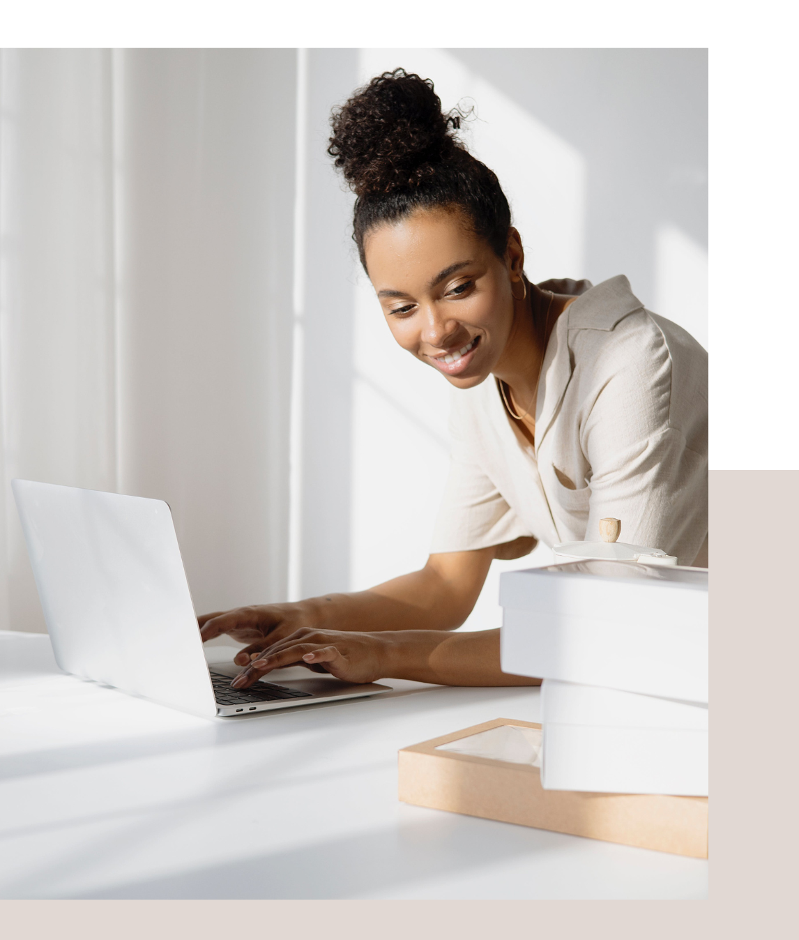 A woman types on a laptop at a white desk with stacked boxes nearby, in a bright room with natural light.